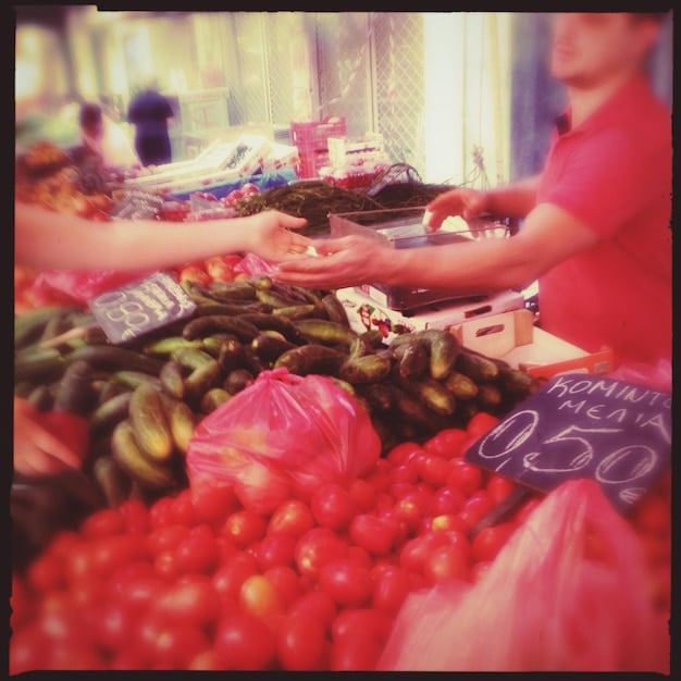 A close-up shot of a local market scene, filled with colorful spices, fruits, and vegetables, with vendors and shoppers interacting in the background.