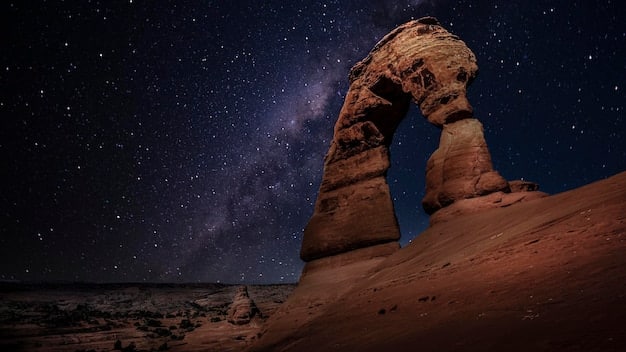 A long-exposure photograph of Delicate Arch in Arches National Park, with the Milky Way visible through the archway. Light painting illuminates the arch from below, creating a warm glow.