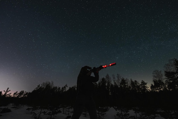 A person setting up a telescope in a dark field, under a sky full of stars. They are wearing a headlamp and using a star chart to locate constellations in the night sky.