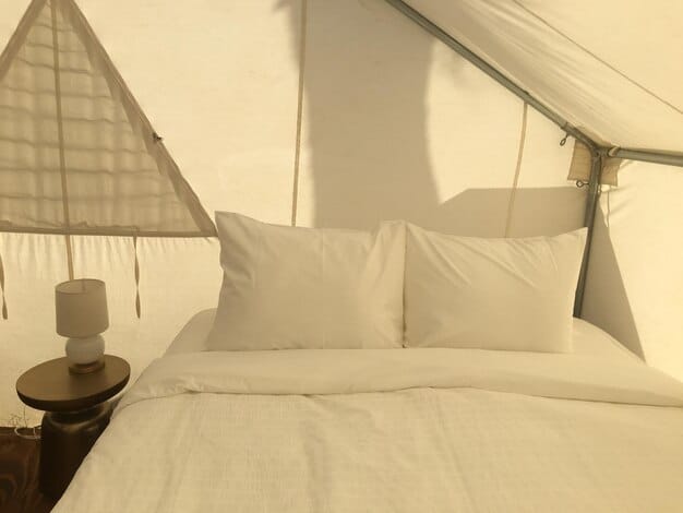 Inside a glamping tent, focus on a king size bed with premium bedding, decorative pillows, and a soft, warm-toned throw blanket. A bedside table features a stylish lamp, books, and a glass of wine. The tent walls are canvas, creating a cozy atmosphere.