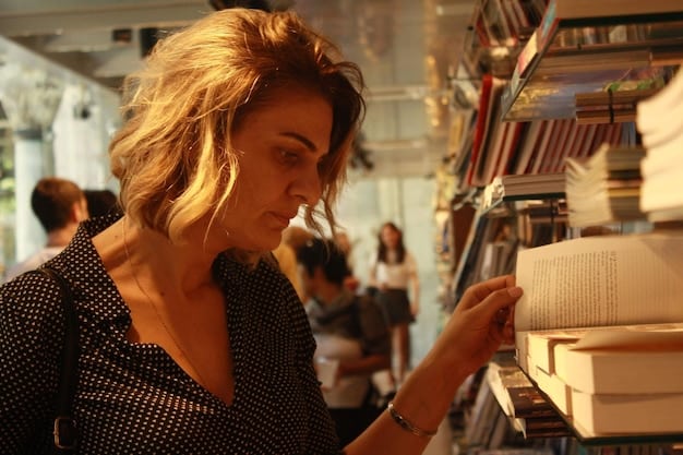 A cozy independent bookstore with floor-to-ceiling shelves filled with books. A person is browsing the shelves, and a local author is giving a reading in the background. The scene captures the intimate and literary atmosphere.